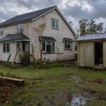 Maison individuelle après une forte tempête, avec un abri de jardin et une serre visibles dans un jardin détrempé sous un ciel venteux.